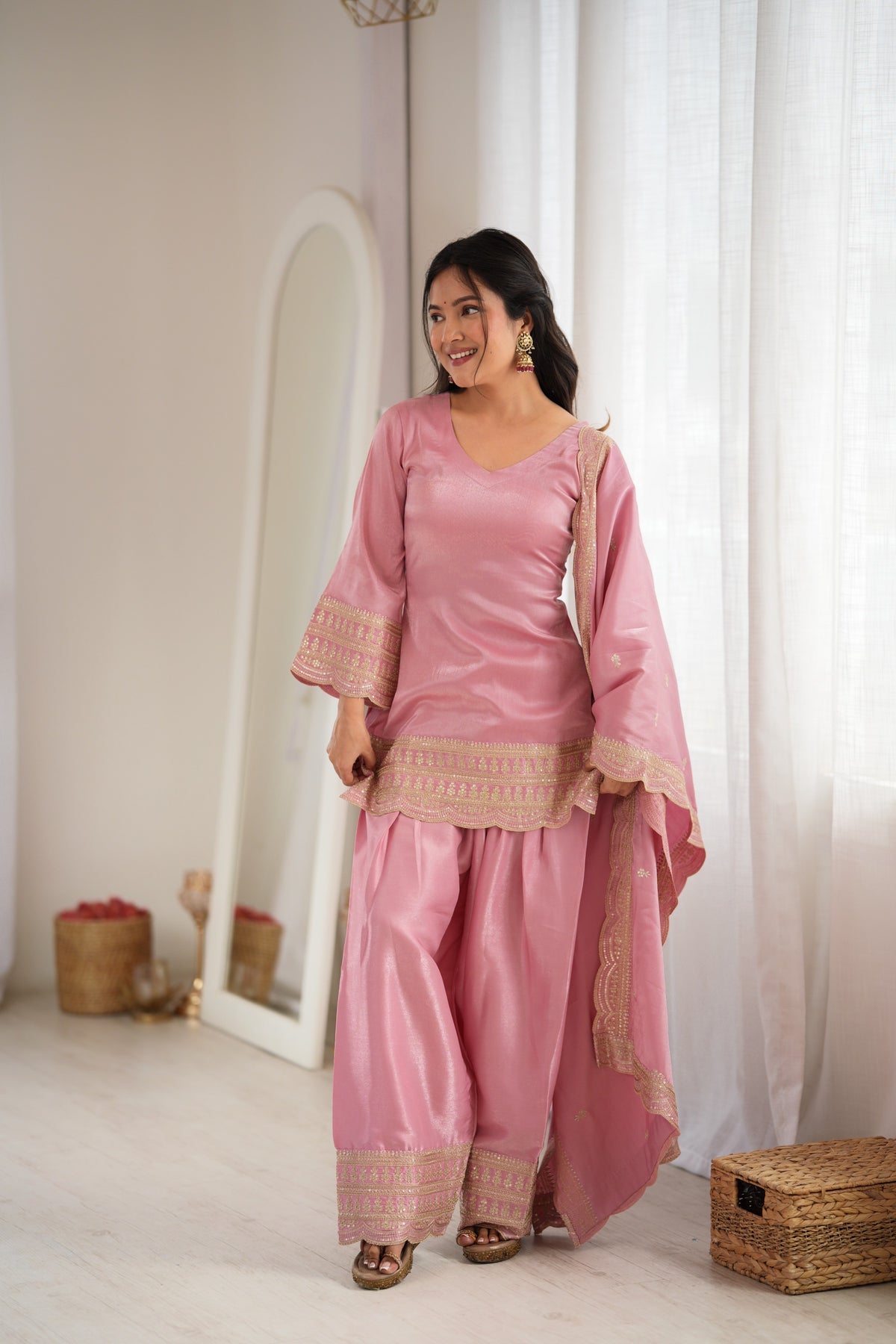 A woman modeling a pink traditional three-piece outfit with beige embroidered borders, standing in a bright, minimalist room with white curtains and a floor mirror.