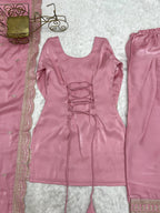 A pink traditional outfit laid flat on a white textured surface, including a lace-up top with long ties, matching pants, and a patterned pink fabric with metallic trim.