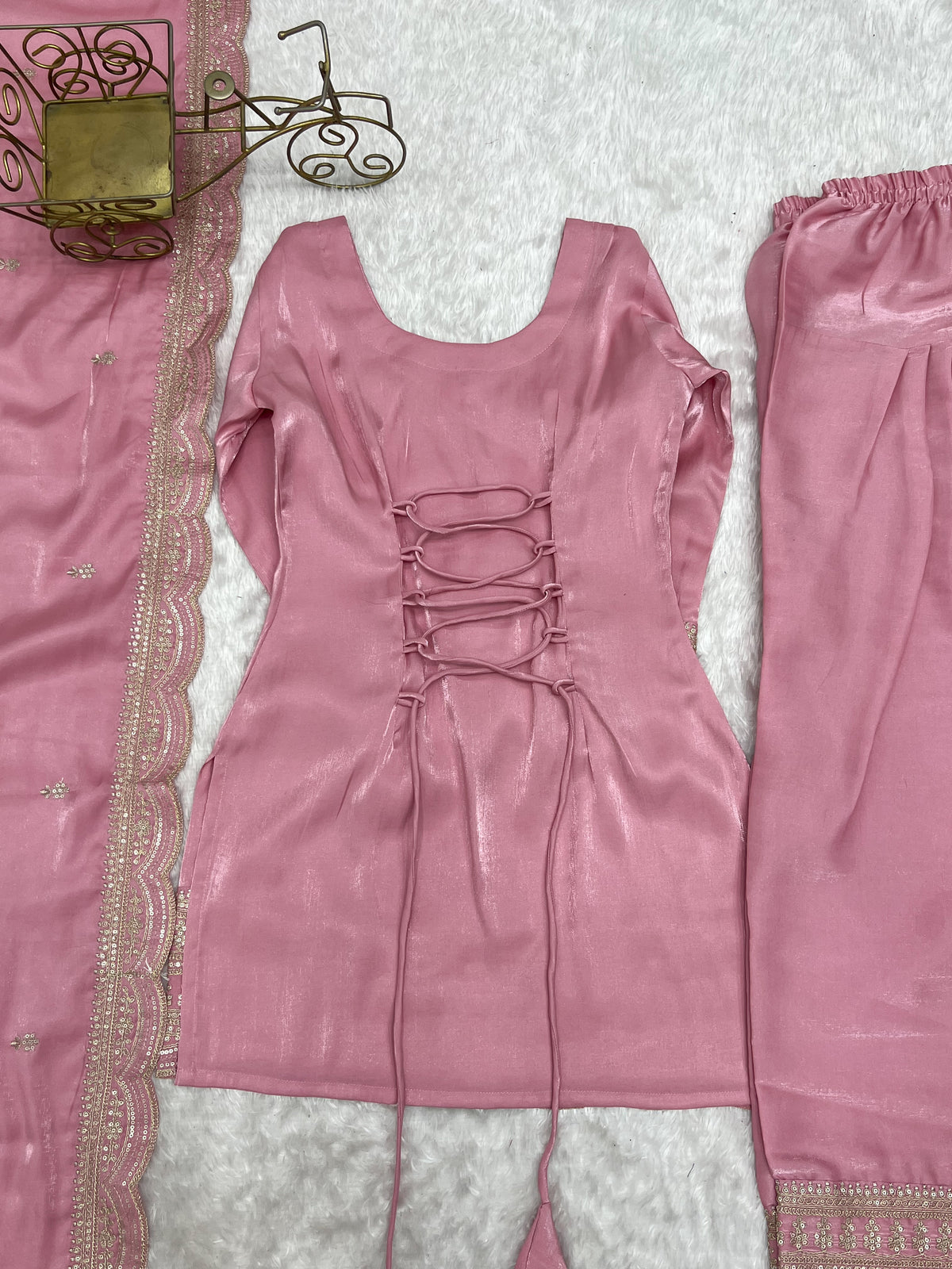 A pink traditional outfit laid flat on a white textured surface, including a lace-up top with long ties, matching pants, and a patterned pink fabric with metallic trim.