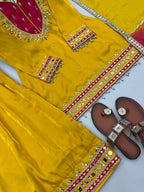 A bright yellow traditional embroidered outfit laid flat on a white surface, including a kurta or dress with a deep red neckline, matching yellow pants, and brown sandals with metallic embellishments.